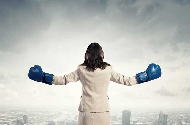 Reverse shot of woman in business suit with arms outstretched, wearing boxing gloves.